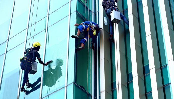 Three window cleaners suspended on ropes clean a modern glass building facade during the day.